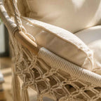 Close-up of a cream macrame hanging chair with thick woven rope, a light wood armrest, and plush cream cushions in a softly lit indoor setting.