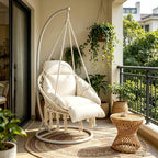 Macrame hanging chair with white cushions on a balcony with plants and a small table.